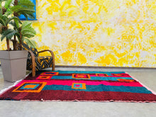 Load image into Gallery viewer, A vibrant, multicolored Moroccan rug laid out in a sunny room with blue window shutters and a leafy plant.

