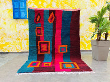 Load image into Gallery viewer, Full view of a Moroccan rug with bold geometric designs, displayed against a yellow wall with a plant and furniture.
