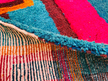 Load image into Gallery viewer, A section of a Moroccan rug showing a close-up of the pattern and fibers, with vibrant reds and blues dominating.
