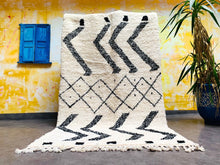 Load image into Gallery viewer, Striking Beni Ourain Moroccan rug with black zigzag and diamond patterns displayed vertically, adding a bold statement to a room with yellow walls and blue window shutters.
