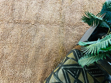 Load image into Gallery viewer, Detailed texture of a high-pile brown Moroccan wool rug, partially draped with green plant leaves and wooden art, emphasizing the cozy and handcrafted quality
