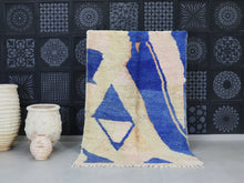 Load image into Gallery viewer, Moroccan wool rug with abstract blue and beige patterns displayed in a cozy setting.
