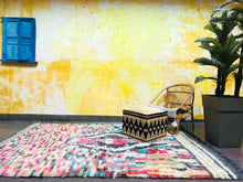 Load image into Gallery viewer, A vibrant Moroccan wool rug with a colorful geometric pattern, displayed in a room with a yellow wall and blue window shutters.
