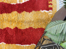 Load image into Gallery viewer, Detailed view of a Moroccan wool rug&#39;s edge, showcasing the transition from dense red and yellow patterns to a white fringe border.
