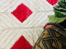 Load image into Gallery viewer, Close-up of a Moroccan Berber rug featuring bold red diamond designs, alongside lush fern leaves and ethnic wooden decor.
