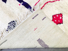 Load image into Gallery viewer, Corner detail of a Moroccan wool rug, with rich purple and vibrant pink accents on a natural wool background, exuding artisanal charm.
